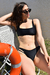 Mujer sentada en una silla de playa, vistiendo un bikini negro, con gafas de sol, junto a un salvavidas y un lago al fondo.