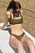 Modelo sentada en la arena usando un bikini verde oliva, con gafas de sol y una toalla blanca debajo.