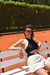 Mujer sentada en una banca, usando un top negro y pantalones cortos claros, con gafas de sol, en una cancha de tenis de polvo de ladrillo.