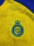 Detalle del escudo del club Al Nassr en la camiseta titular de Cristiano Ronaldo, con fondo amarillo y azul.