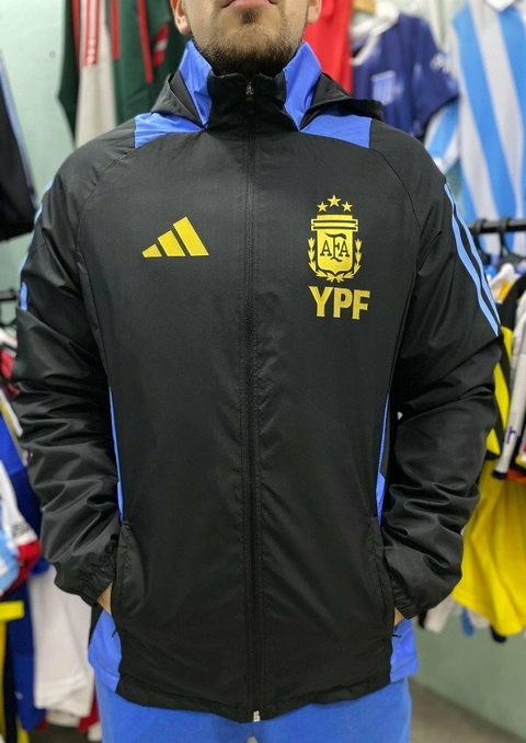 Hombre usando un rompeviento negro con detalles en azul y el logo de la AFA y YPF en el pecho.