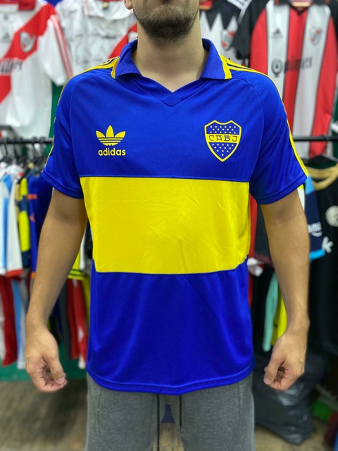Camiseta de Boca Juniors con colores azul y amarillo, modelo reedición, vista frontal en un fondo de otras camisetas.