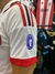 Camiseta blanca de fútbol con detalles en rojo y negro, mostrando el escudo de un torneo en la manga.