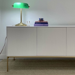 Mueble consola blanco con puertas varilladas, adornado con una lámpara verde y objetos decorativos como libros y una taza.