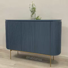 Mueble tipo aparador de color azul con diseño de listones verticales y patas doradas, decorado con un jarrón y flores en la parte superior.