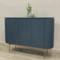Mueble de diseño moderno en color azul con puertas acanaladas y patas doradas, decorado con un jarrón y flores.