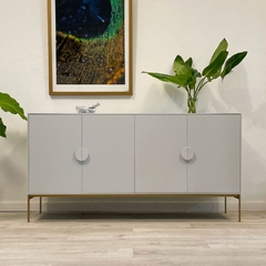 Vista de frente del mueble vajillero, con líneas rectas y limpias, y espacios amplios. Posee 4 puertas con sistema push, y estante regulable en el interior. Su color es un blanco nube delicado y suave, que combina a la perfección con su base dorada metáli
