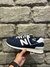 Tenis New Balance 574V2 na cor azul marinho, visto de lado, com detalhes em branco, contra um fundo de parede de pedra.