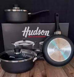 Conjunto de cacerolas antiadherentes de la marca Hudson, incluyendo una olla con tapa y una sartén, presentado sobre una caja con el logo de la marca.