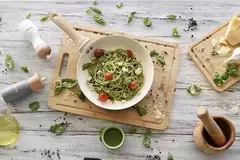 Plato de pasta con salsa verde y tomates cherry en una sartén de cerámica crema, sobre una tabla de madera con ingredientes frescos alrededor.