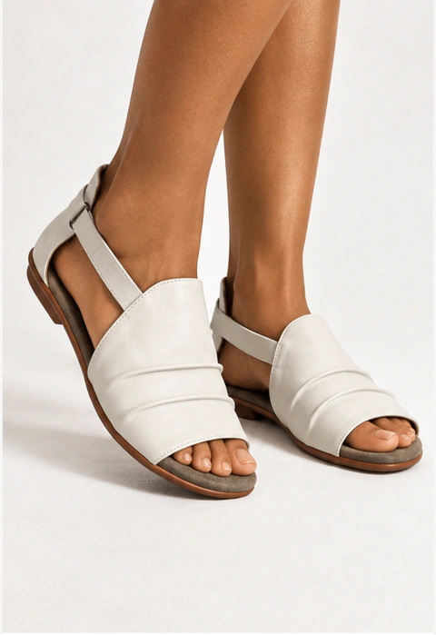 Sandália feminina rasteira em couro legítimo off-white. O modelo apresenta cabedal largo com detalhes plissados em pregas transversais, abertura lateral e calcanhar aberto com tira fina de fechamento ajustável. A palmilha é em camurça cinza e o solado pla