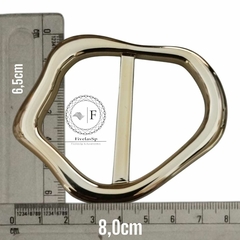 Fivela metal dourada modelo orgânica, medindo 6,5 cm por 8 cm, sobre uma régua.