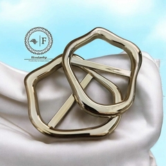 Fivela metal dourado modelo orgânico com reforço, mostrando duas peças em cima de um tecido claro.