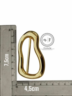 Fivela metálica dourada em formato orgânico, medindo 7,5 cm de altura e 4,5 cm de largura, acompanhada de régua para referência de tamanho.