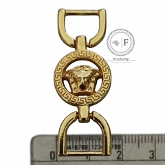 Fivela de metal dourado com design de medalhão, mostrando uma cabeça de leão, em uma régua para referência de tamanho.