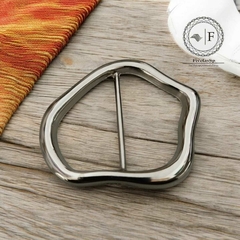 Fivela orgânica de metal na cor prata, com design irregular, sobre uma superfície de madeira clara.