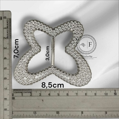 Fivela de plástico modelo borboleta na cor prata, com dimensões de 7 cm de altura, 8,5 cm de largura e 3 cm de espessura, ao lado de uma régua para referência de tamanho.