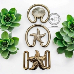 Fivelas plásticas douradas em diferentes modelos, apresentando um design de concha e estrela do mar, dispostas ao lado de suculentas verdes.