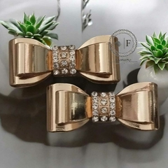 Dois laços em metal dourado com strass e detalhe em garra, exibidos sobre superfície espelhada com plantas decorativas ao fundo