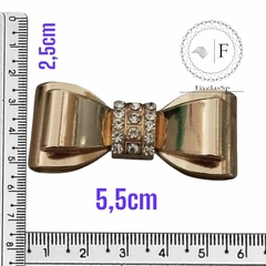 Laço em metal dourado com strass, medindo 5,5 cm de largura por 2,5 cm de altura, com régua ao redor para referência de tamanho