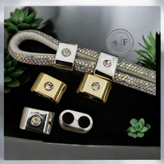 Passador enforcador regulador de plástico com strass em diferentes cores, incluindo peças douradas e prateadas, apresentadas sobre um fundo escuro com plantas.