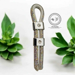 Passador enforcador regulador de plástico com strass, exibindo detalhes brilhantes e um design elegante, sobre fundo com plantas verdes.