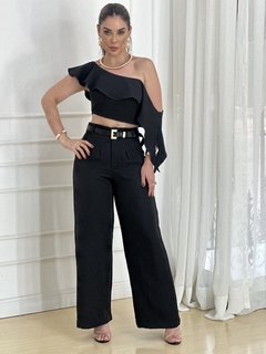 Modelo vestindo um conjunto preto com cropped assimétrico e calça alfaiataria, posando em um ambiente iluminado.
