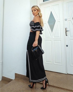 Modelo usando um conjunto de top com babados e saia midi, na cor preta com detalhes em branco, segurando uma bolsa preta.