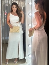 Modelo vestindo um vestido branco longo com amarração na cintura, segurando uma bolsa de palha e posando em frente a um espelho com luzes.