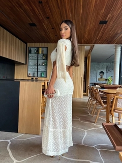 Modelo usando um vestido longo branco de renda, com detalhes em franjas, em um ambiente moderno e sofisticado.