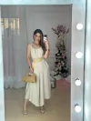 Mulher posando com macacão Jasmine Alça Dupla, acessórios dourados e bolsa de palha em frente a um espelho.