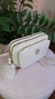 Bolsa porta celular com pompom na cor branca, com detalhes em dourado e alça de ombro, colocada sobre um puff rosa e decorada com folhas verdes ao fundo.