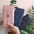 Três carteiras de mão grandes em cores rosa, azul e preta, com detalhes de botão decorativo, sendo seguradas por uma mão, com fundo de plantas verdes.
