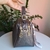 Bolsa baú média marrom com alça de mão, detalhes em metal dourado e tassel cinza pendurado