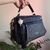 Bolsa baú média preta com alça de mão, detalhe de pingente com tassel preto e pedra branca, sendo segurada por uma mão, fundo com plantas