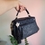 Bolsa baú média preta com alça de mão, detalhe de tassel pendurado, sendo segurada por uma mão feminina