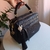 Bolsa baú média preta com alça de mão em couro sintético, detalhes dourados e pingente decorativo com tassel preto