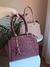 Imagem de uma bolsa feminina grande de ombro com alça trançada, na cor bordô, com um chaveiro em forma de urso; ao fundo, uma bolsa na cor nude.