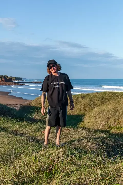 Hombre con remera boxy fit negra Captain Fin Lev y gorra negra, posando en un paisaje costero con pasto, playa y mar de fondo