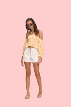 Modelo vestindo shorts brancos e blusa amarela, posando de forma elegante.