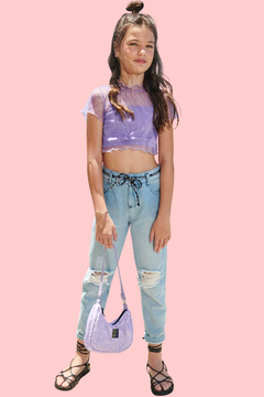 Menina usando calça mom jeans de lavagem clara, blusa cropped roxa e bolsa lilás, posando em um ambiente ao ar livre com vista para o mar.