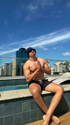 Homem usando um chapéu bucket de crochê preto, sentado à beira de uma piscina, com o céu azul ao fundo.