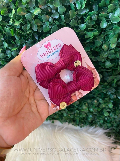 Par de laços personalizados marsala com acabamento em fita de gorgurão, apresentados em cartão da marca Universo da Laceira, segurados por uma mão feminina, com fundo de plantas verdes.