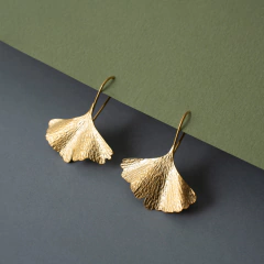 brincos de anzol, feitos a mão em prata 950 e banho de ouro, em formato de folhas de ginkgo biloba da marca Carlos Herrera Joalheria sobre fundo cinza e verde