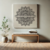 Quadro decorativo com desenho de mandala floral em destaque, adornando uma parede neutra ao lado de uma planta e um banco de madeira.