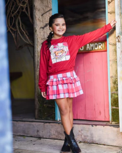 Menina sorridente vestindo conjunto infantil Pink Farm com blusa vermelha estampada de ursinho boiadeiro e saia xadrez rosa e branco, usando botas