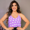Mulher sorridente usando uma blusa roxa com bolinhas brancas, posando com as mãos na cintura.