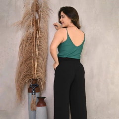 Mulher de costas usando blusa verde e calça preta, posando ao lado de plantas decorativas em um fundo neutro.