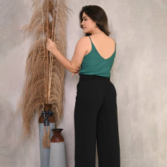 Mulher de costas usando blusa verde e calça preta, segurando plantas decorativas em um ambiente neutro.