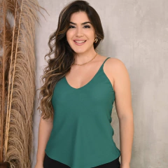 Mulher sorridente usando uma blusa verde com alças finas, em frente a uma parede de textura clara.
